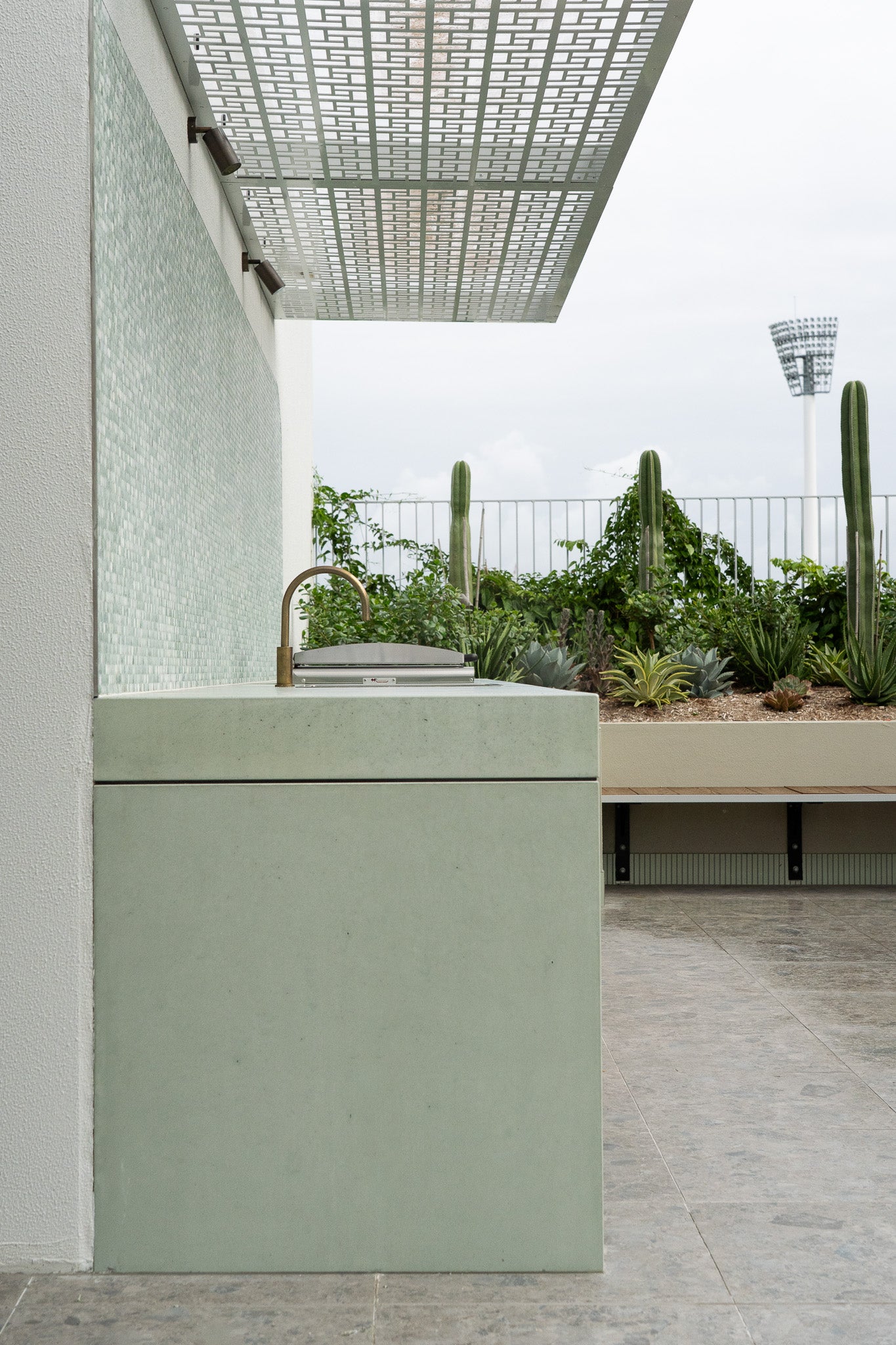 A sleek custom concrete outdoor kitchen unit in a soft sage green finish, featuring an integrated brass tap and compact grill, set against a tiled wall and surrounded by lush rooftop landscaping with cactus and succulents.