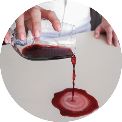 A hand pouring a glass of red whine onto a white concrete benchtop. 
