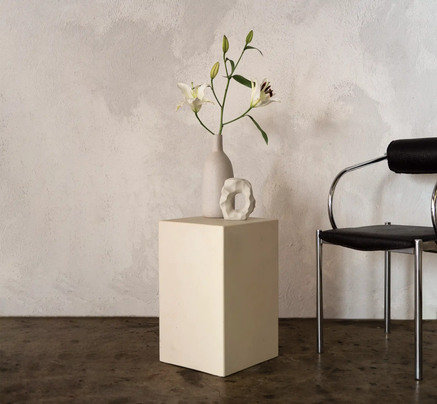 A Sand coloured Sahara Concrete Side Table in a minimalist living room next to a funky modern dining chair and a vase of lilies.