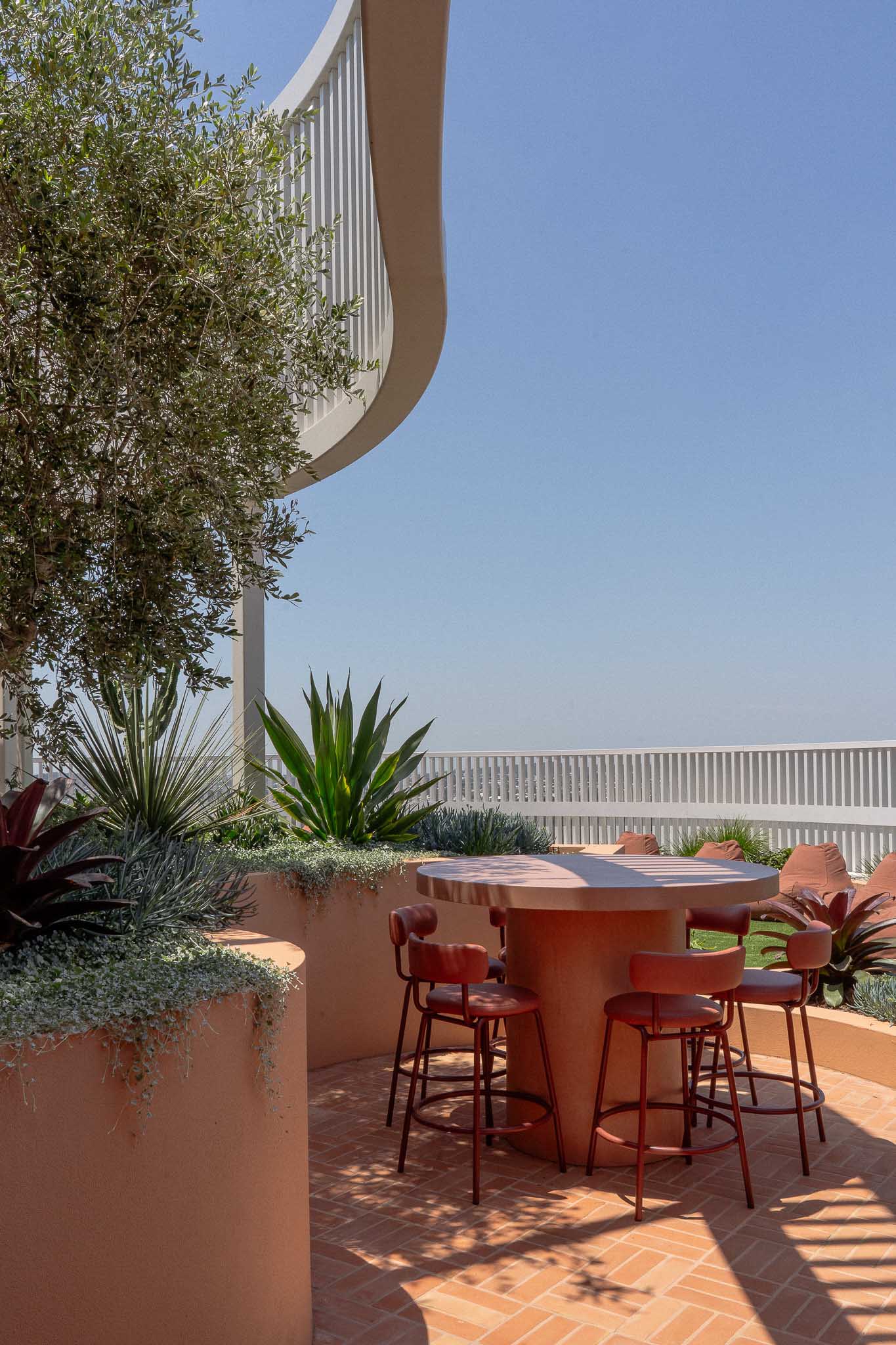 A stylish outdoor seating area featuring a custom round concrete table with a cylindrical base, surrounded by red bar stools with curved backrests. The space is enclosed by terracotta-toned planters filled with lush greenery, including succulents and an olive tree.