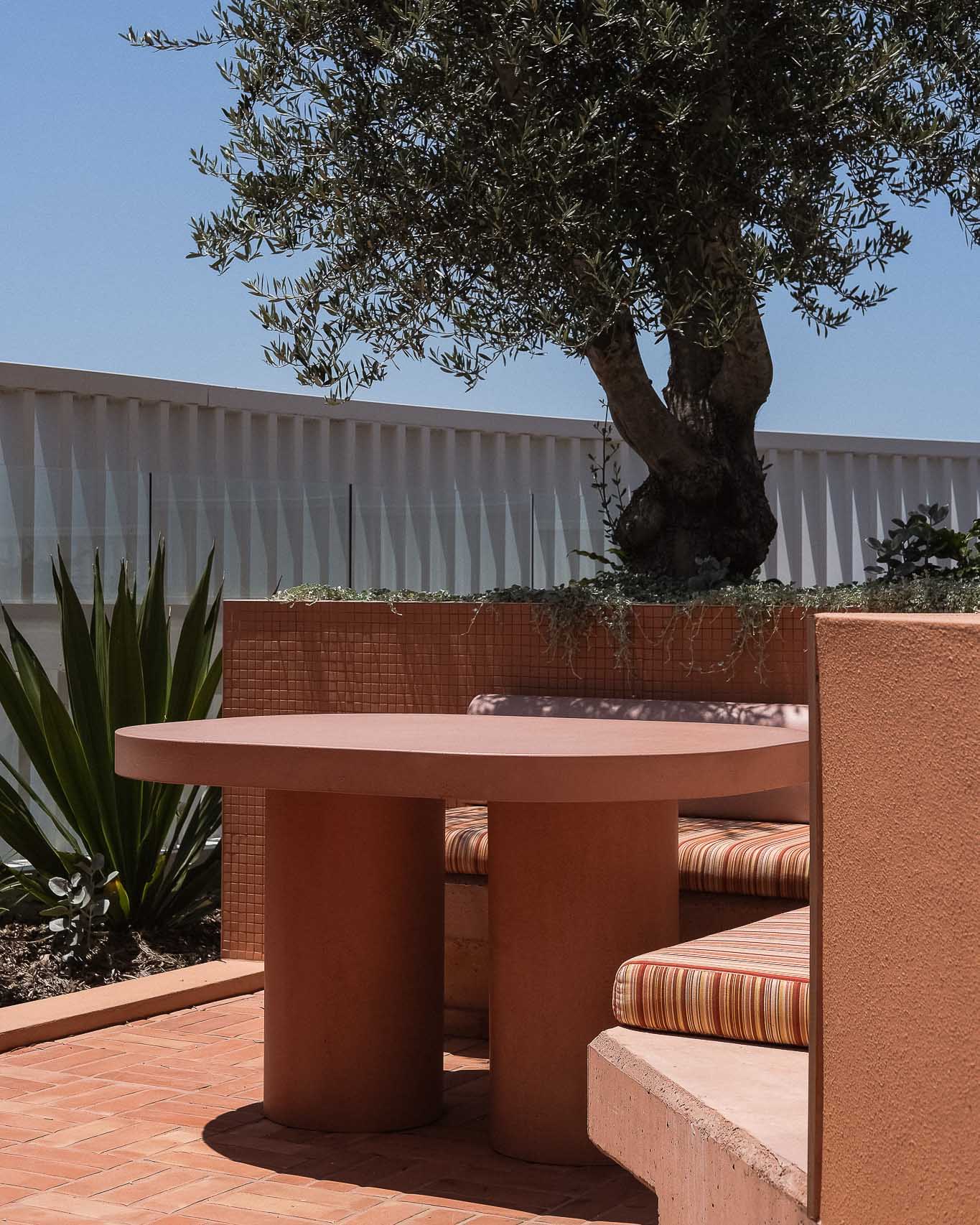 An inviting outdoor dining area featuring a sculptural round concrete table with two cylindrical bases, surrounded by a built-in cushioned bench with striped upholstery in warm tones. The space is framed by terracotta-tiled walls and lush greenery, including an olive tree that provides natural shade. The textured elements, earthy colour palette, and mix of materials create a harmonious and relaxed setting.