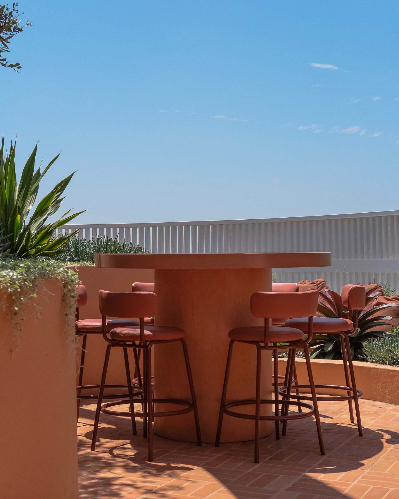 A stylish outdoor seating area featuring a custom round concrete table with a cylindrical base, surrounded by red bar stools with curved backrests.