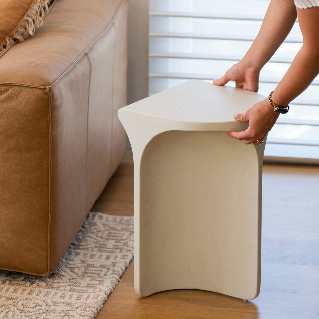 A person lifting the Isle Concrete Side Table, a sculptural and modern piece with a smooth, off-white finish.