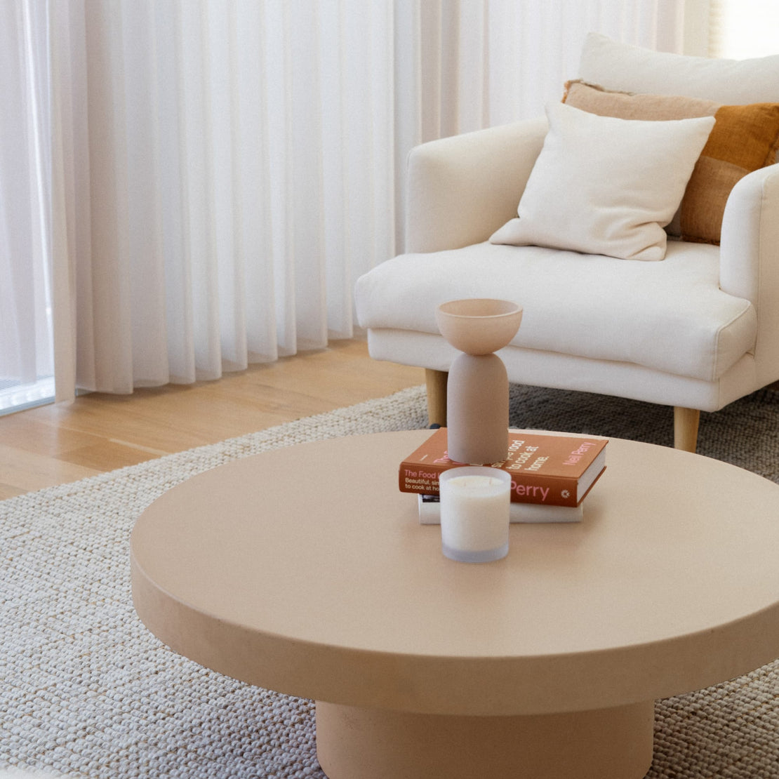 The Siena Concrete Coffee Table in Bisque in a bright white living room. A candle, vase and books sit upon the table.