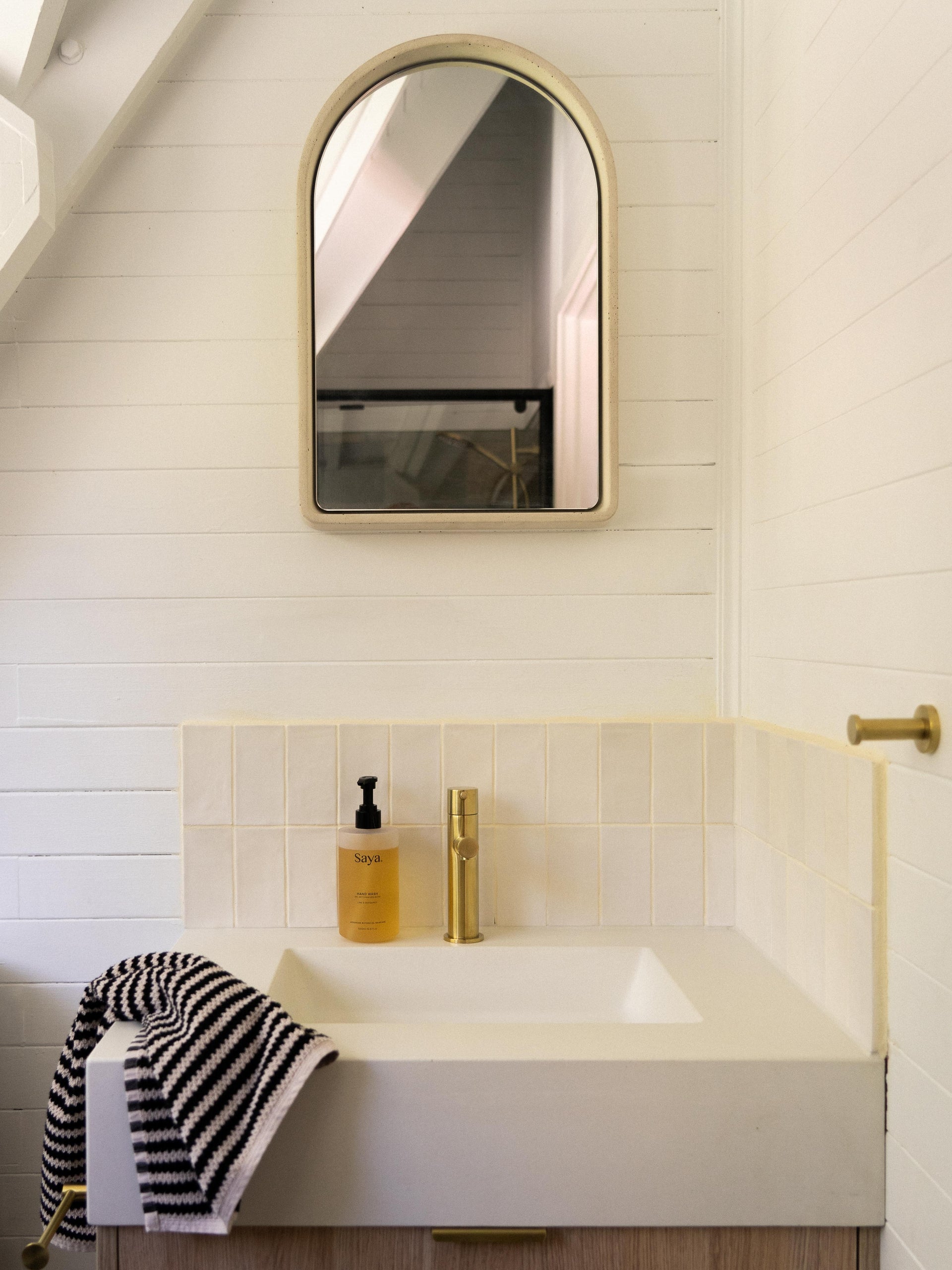 A compact bathroom vanity featuring a minimalist custom concrete basin with soft edges and a slim brass mixer tap. Above it hangs a Arc Concrete Mirror, reflecting the sloped ceiling and black shower frame behind.