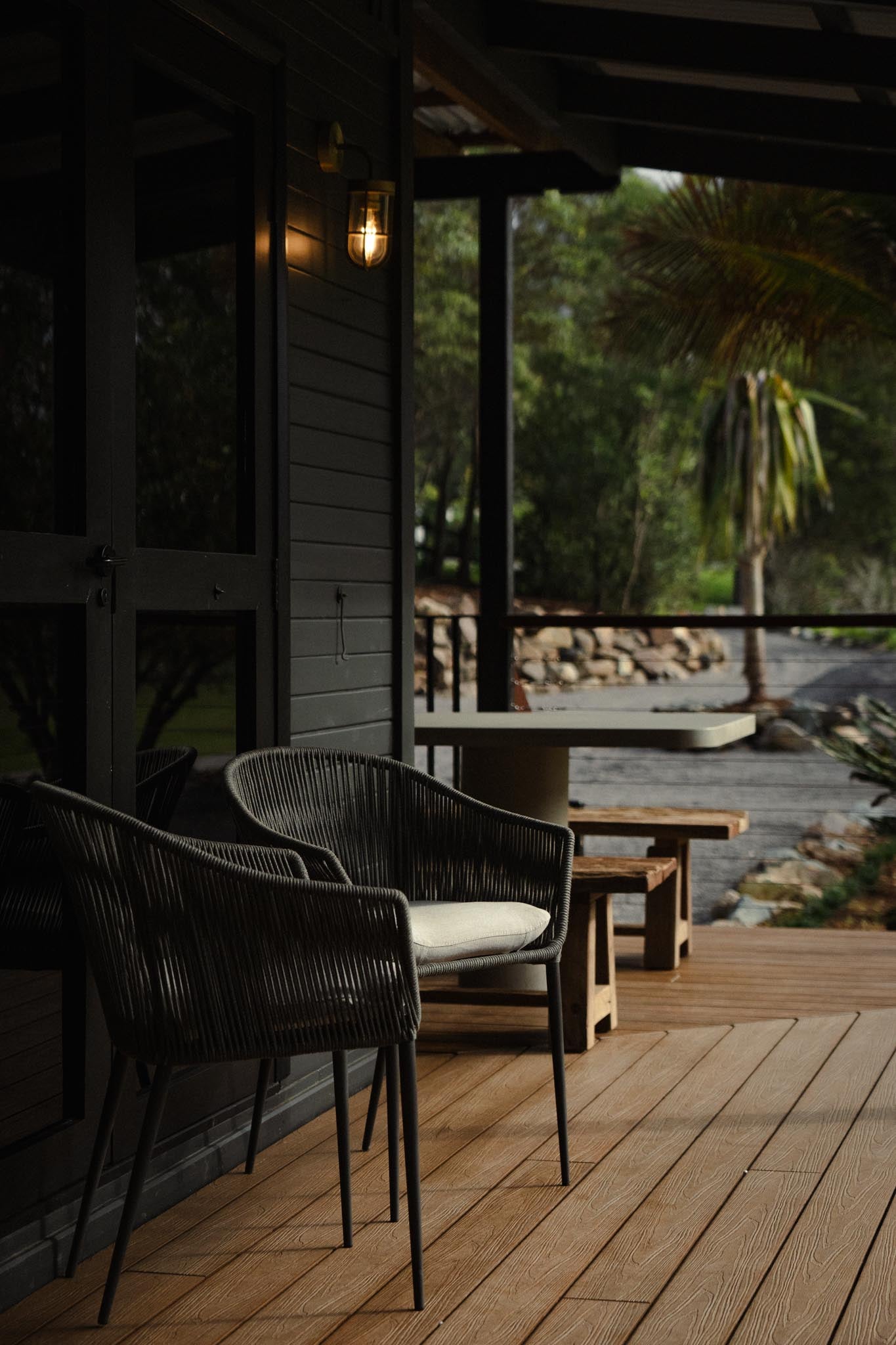 A moody outdoor deck scene at dusk, featuring two black woven lounge chairs with light cushions positioned outside a dark timber-clad building.