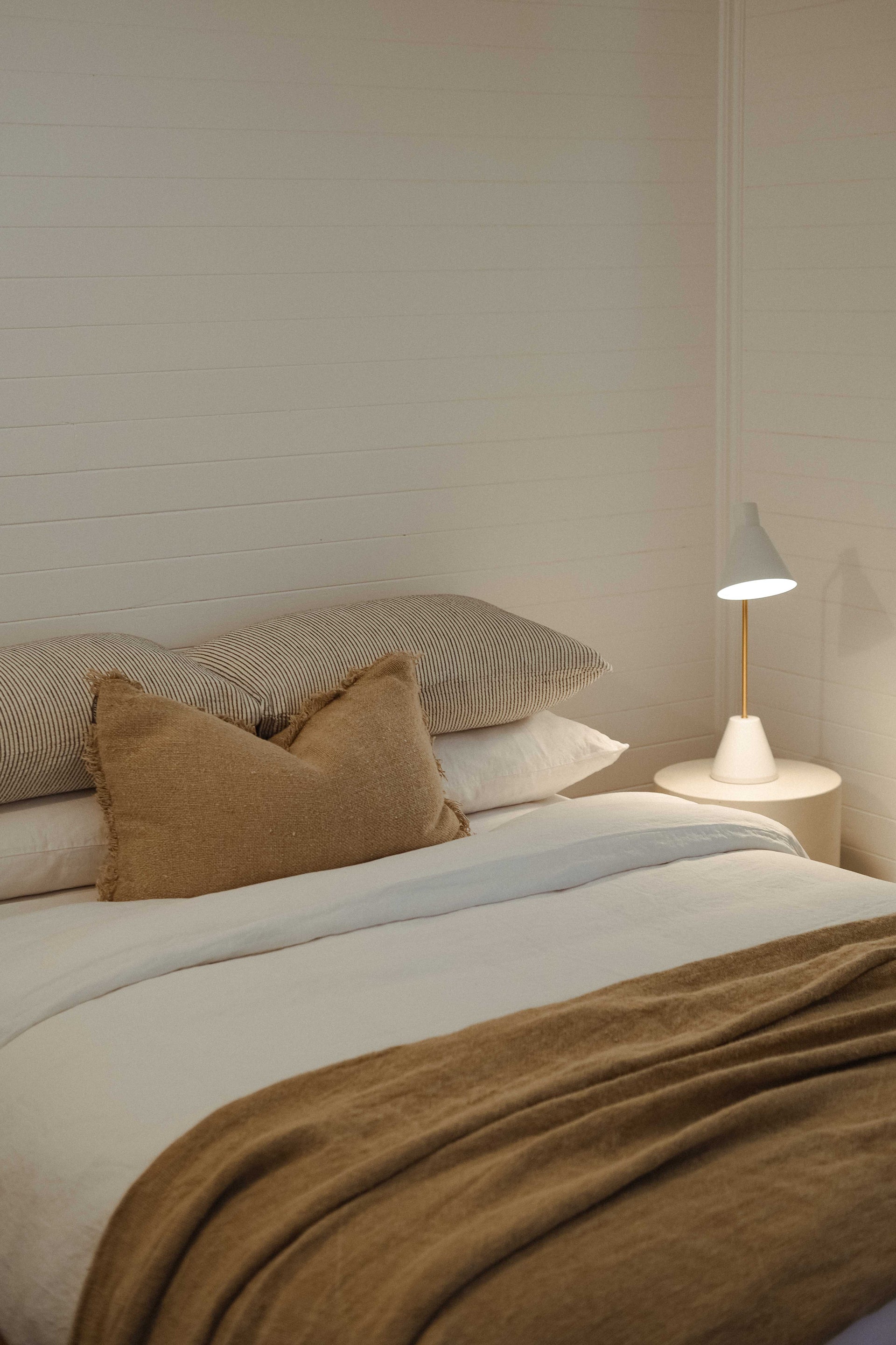 A calming bedroom scene featuring a neatly made bed with white linen, striped pillows, and a textured tan cushion. A soft brown throw is draped across the foot of the bed.