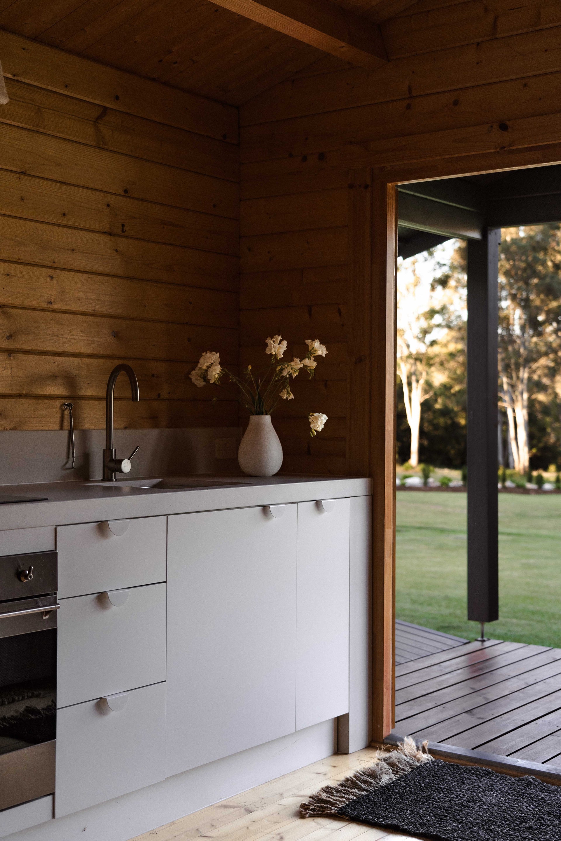 A compact kitchen with matte white cabinetry and a seamless concrete benchtop and splashback, set against warm timber-clad walls and ceiling.
