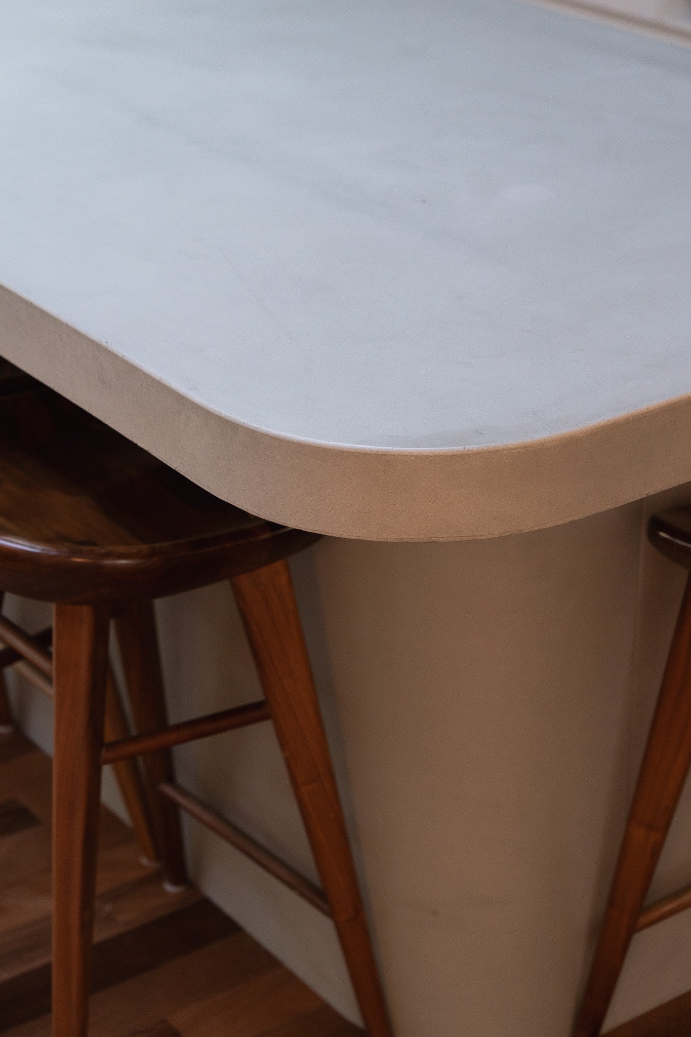 A close-up of a custom concrete kitchen bench with a softly curved edge and smooth finish, paired with timber bar stools on a warm wooden floor, showcasing craftsmanship and refined detailing.