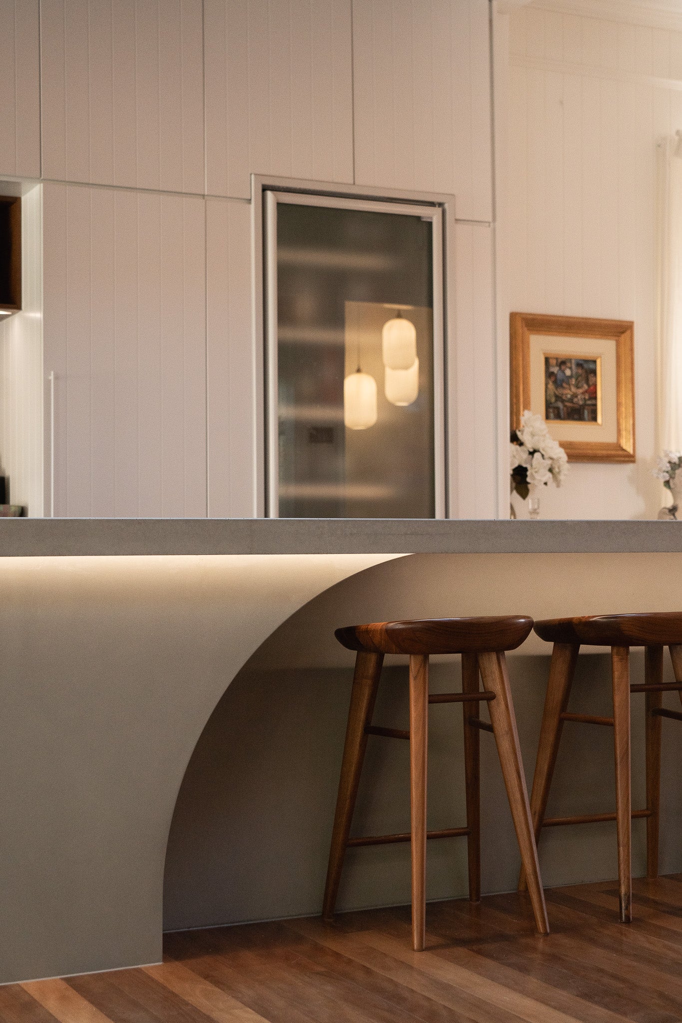  A close-up view of a custom concrete kitchen island with an elegant curved base and under-bench lighting, paired with timber bar stools, set against a white panelled wall with soft pendant lighting and framed artwork in the background.