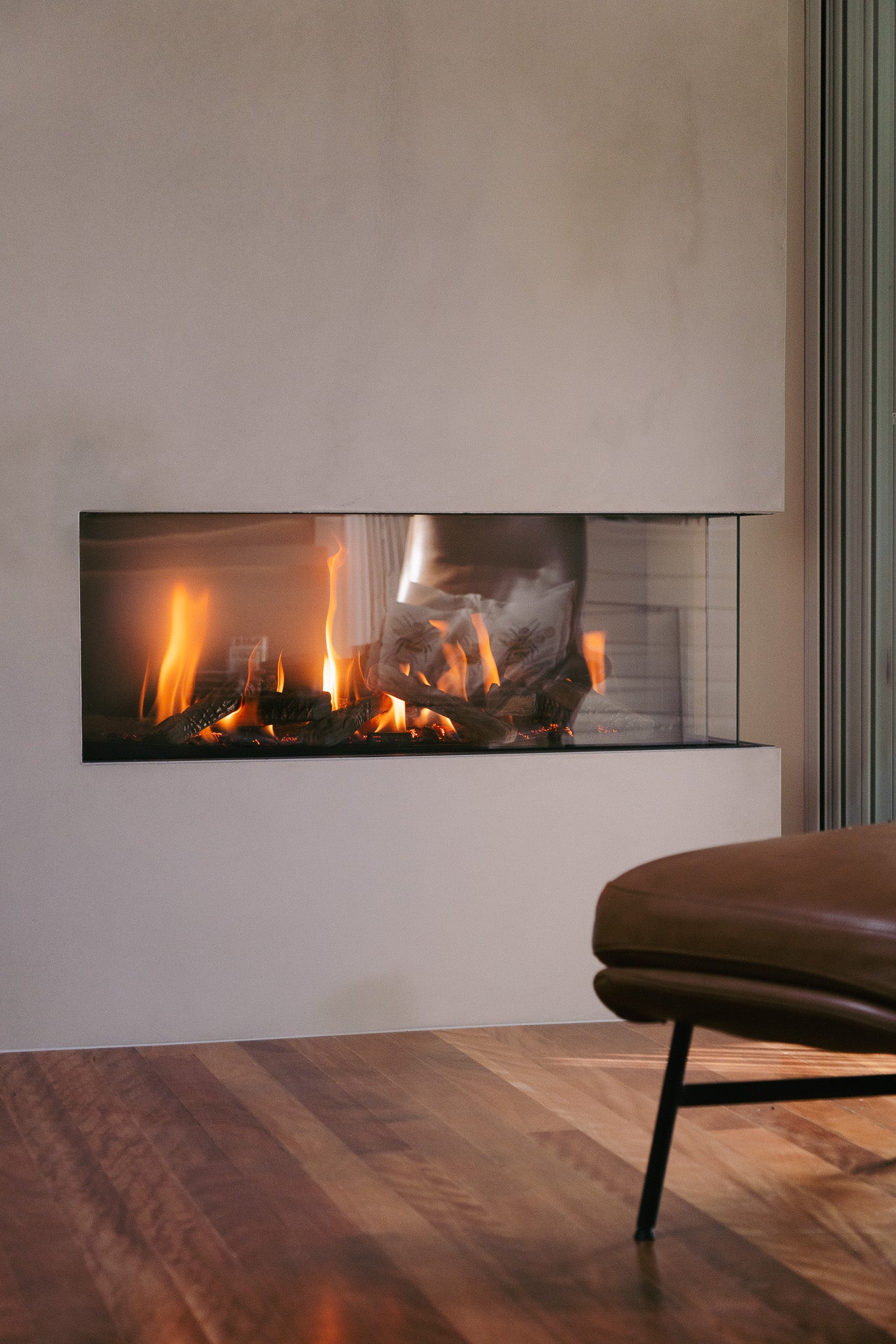 A sleek built-in gas fireplace with a custom concrete surround and glass front, set against a timber floor with a leather lounge chair in the foreground, creating a warm and contemporary living space.
