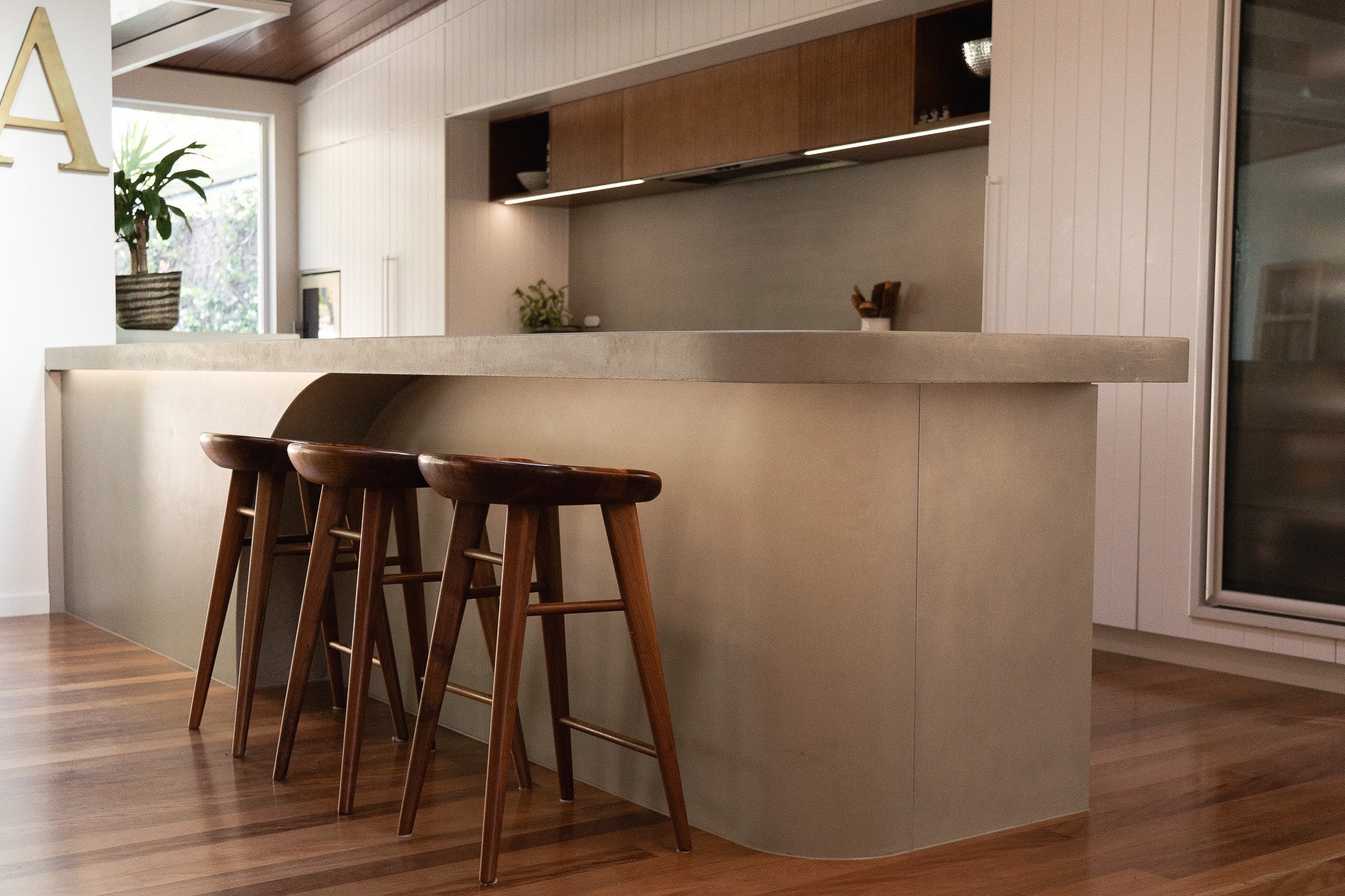 A full view of a custom concrete kitchen island with a soft curved profile and under-bench lighting, complemented by timber bar stools and set within a contemporary kitchen featuring warm wood flooring, white panelling, and timber cabinetry.