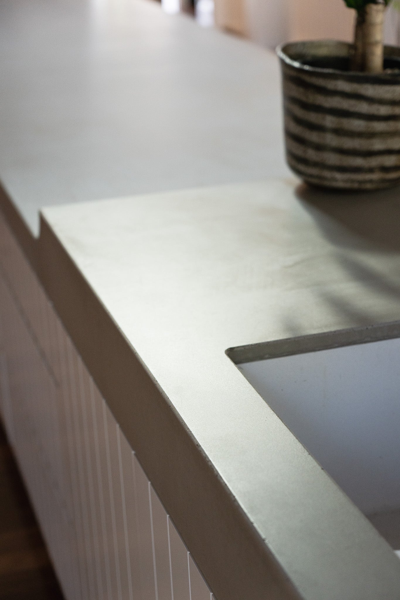 A detail view of a custom concrete kitchen island with a seamless integrated sink and smooth matte finish, paired with vertical panel cabinetry and soft natural lighting.