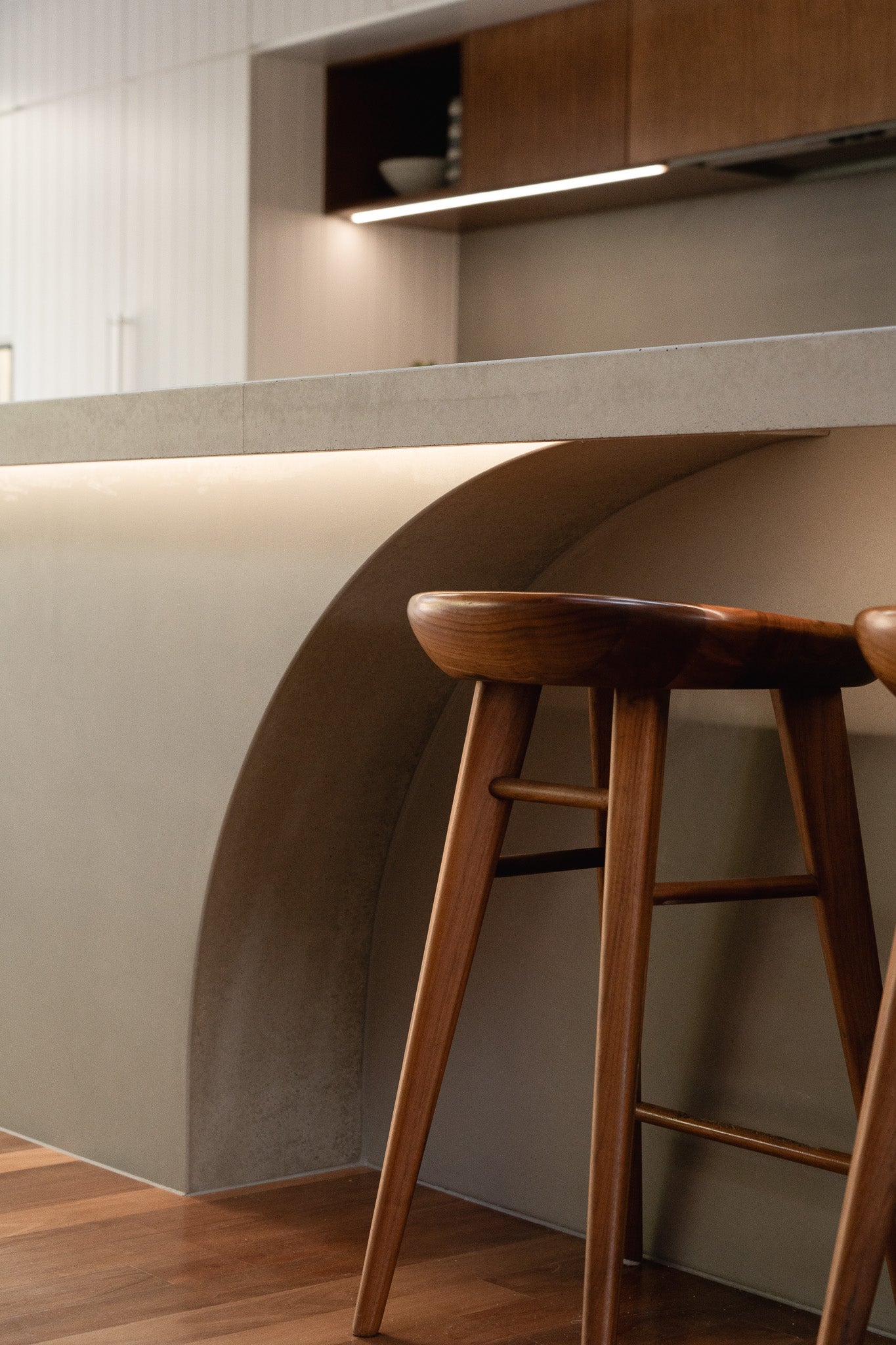 A custom concrete kitchen island with a softly curved arch detail and integrated LED lighting, paired with sculpted timber bar stools and warm timber flooring, creating a refined and inviting atmosphere.