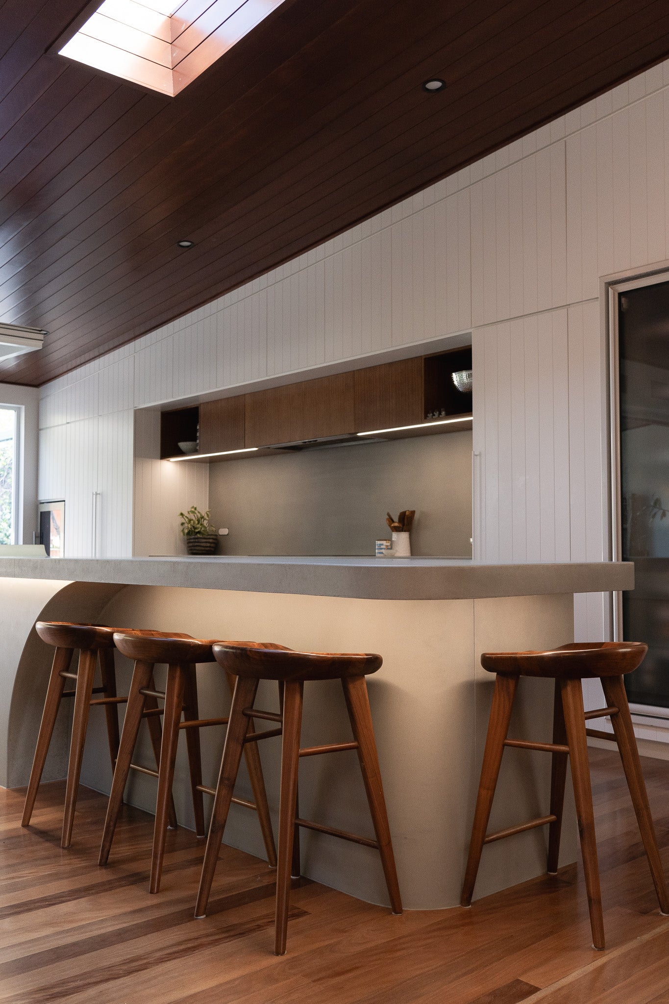  A spacious modern kitchen featuring a custom concrete island with curved detailing and under-bench lighting, surrounded by timber bar stools and warm wood flooring, set beneath a rich timber ceiling with a skylight.