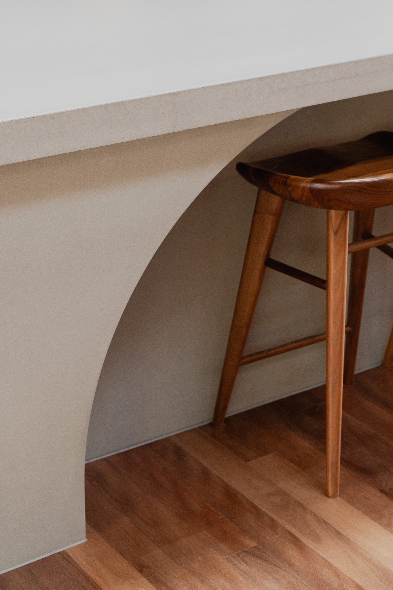 A close up down angle of kitchen featuring a custom concrete island with curved detailing and under-bench lighting, surrounded by timber bar stools and warm wood flooring.