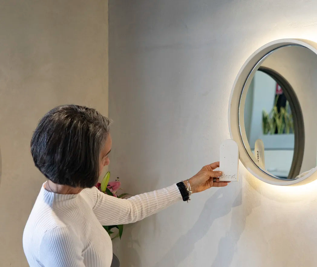 A woman holding a concrete sample up to a round Sol Concrete Mirror in the Blend Concrete Design Showroom.