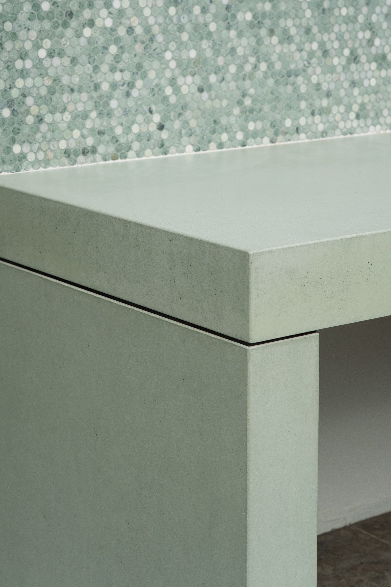Close-up of a custom pale green concrete bench with clean mitred edges and a sharp corner detail, set against a textured mosaic tile splashback in soft green and white tones.