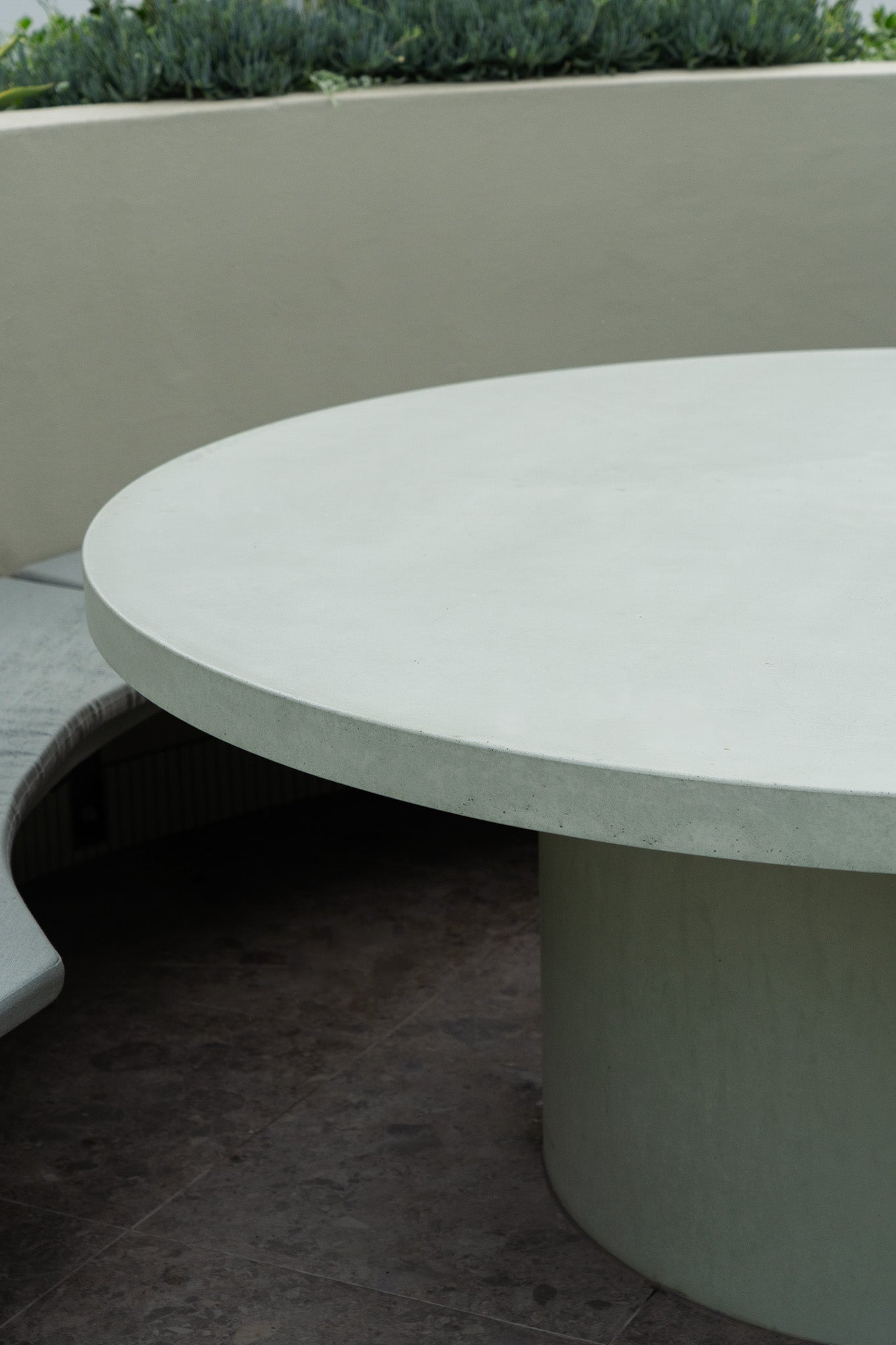 Close-up of a round green concrete table with a solid cylindrical base, positioned next to curved built-in bench seating in an outdoor setting with soft landscaping and stone flooring.