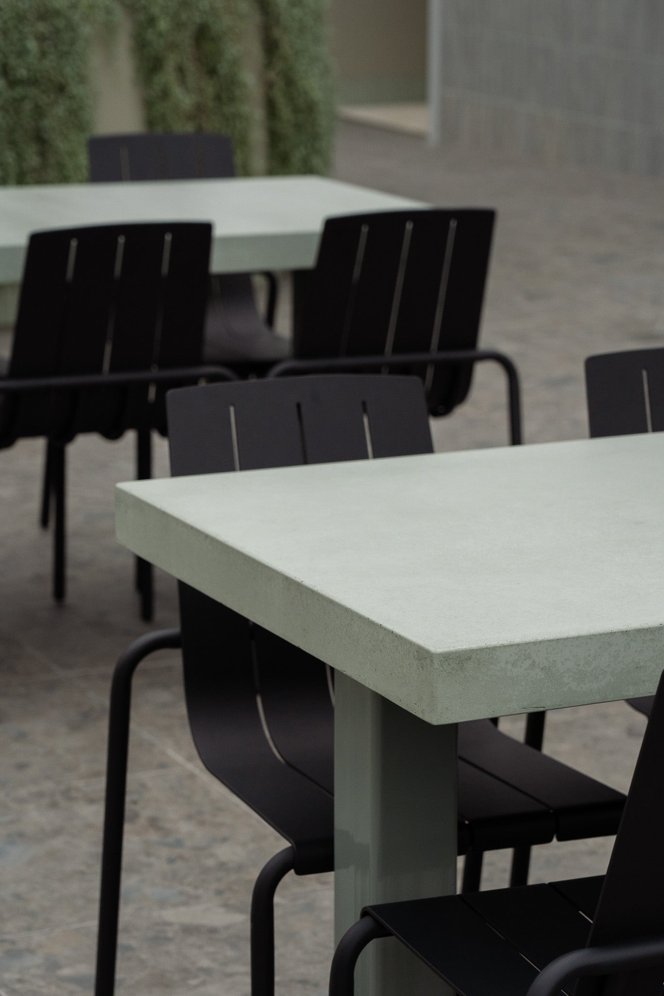 Detail view of a rectangular green concrete outdoor dining table with a thick, chamfered edge and metal pedestal base, surrounded by black metal chairs. 