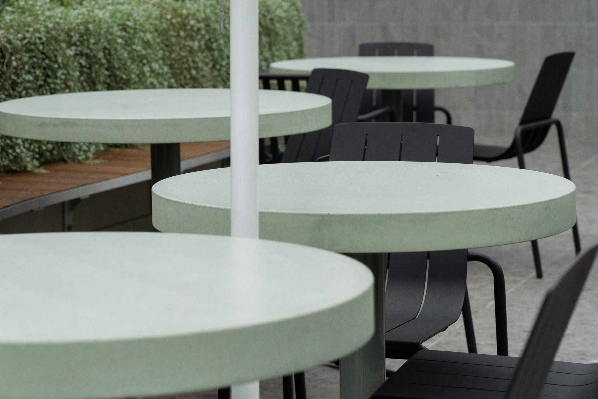 A cluster of custom round concrete tables in a soft sage green finish, positioned on a modern rooftop space with dark outdoor chairs and a backdrop of lush greenery and tiled walls.