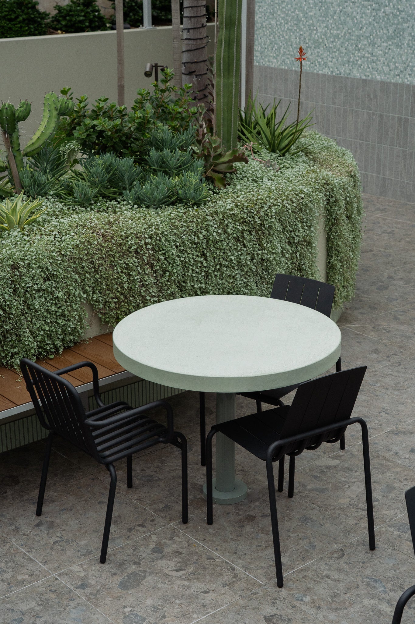 A custom round green concrete table with a central pedestal base, paired with three black outdoor chairs on a tiled surface. The setting features lush greenery, cascading plants, and a timber bench integrated into a curved raised garden bed.