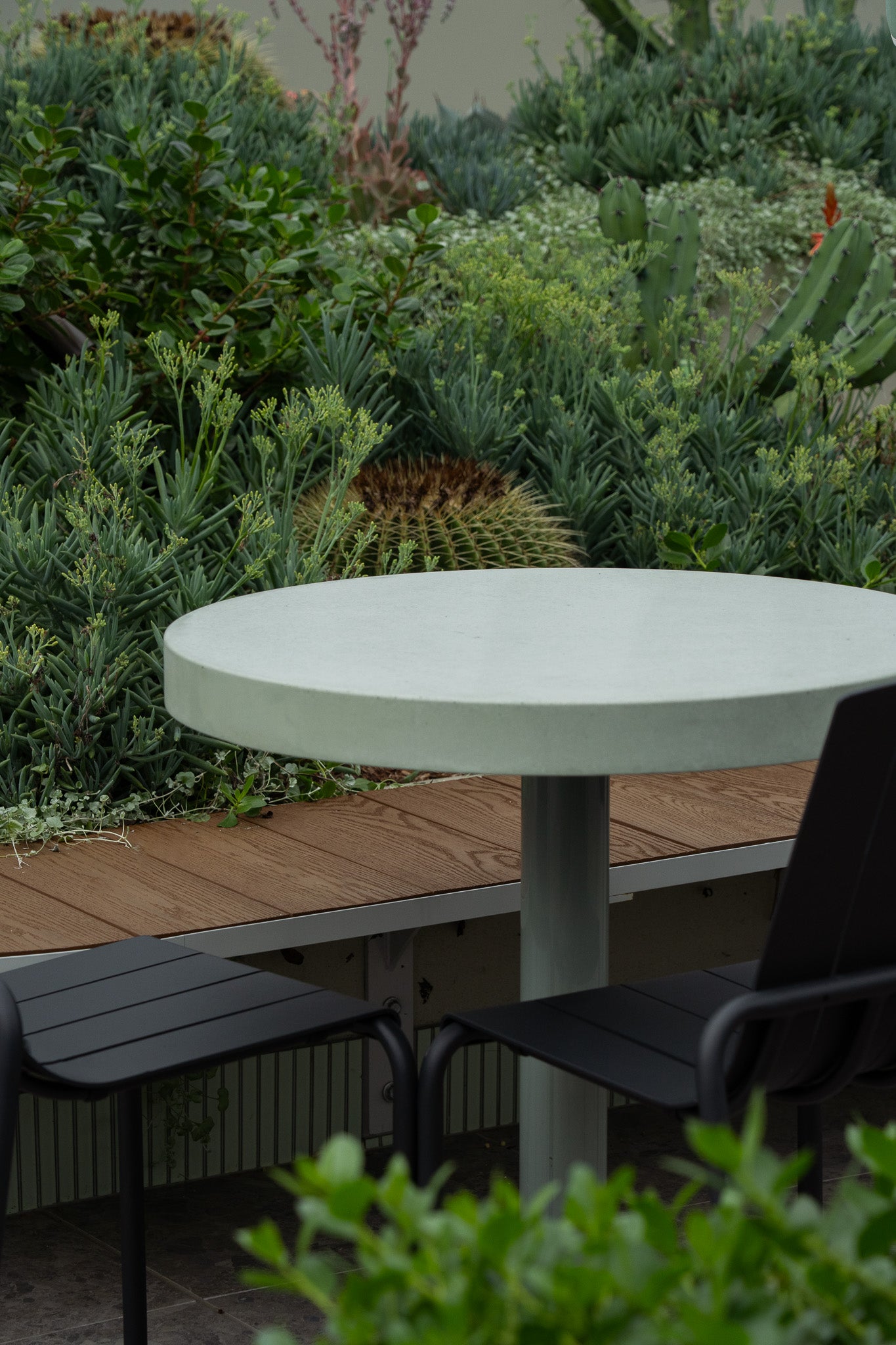 A custom round green concrete table with a central pedestal base, paired with three black outdoor chairs on a tiled surface. The setting features lush greenery, cascading plants, and a timber bench integrated into a curved raised garden bed.