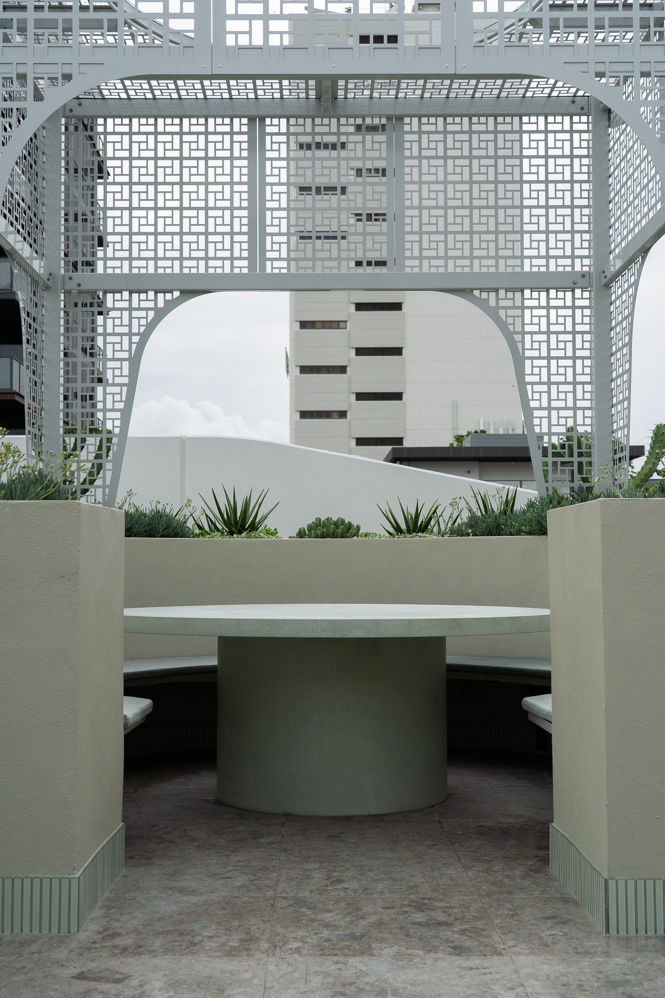 A large round green concrete table with a solid pedestal base, set within a curved built-in bench seating area. Surrounded by soft green planters and a geometric metal pergola structure, the space overlooks an urban cityscape in the background.
