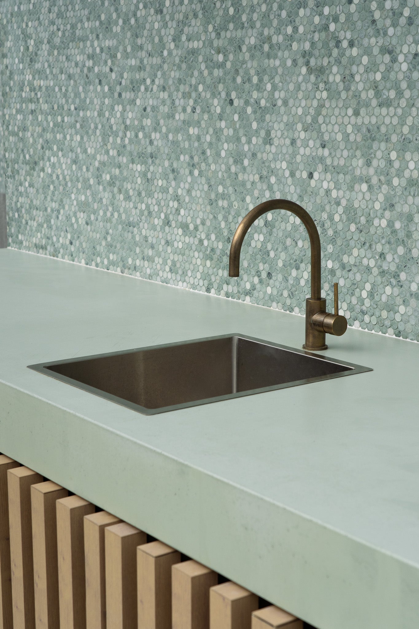 A pale green custom concrete benchtop with an integrated stainless steel sink and brushed brass tap, set against a mosaic tile splashback in soft green and white tones. The cabinetry features vertical timber slats for added texture and warmth.