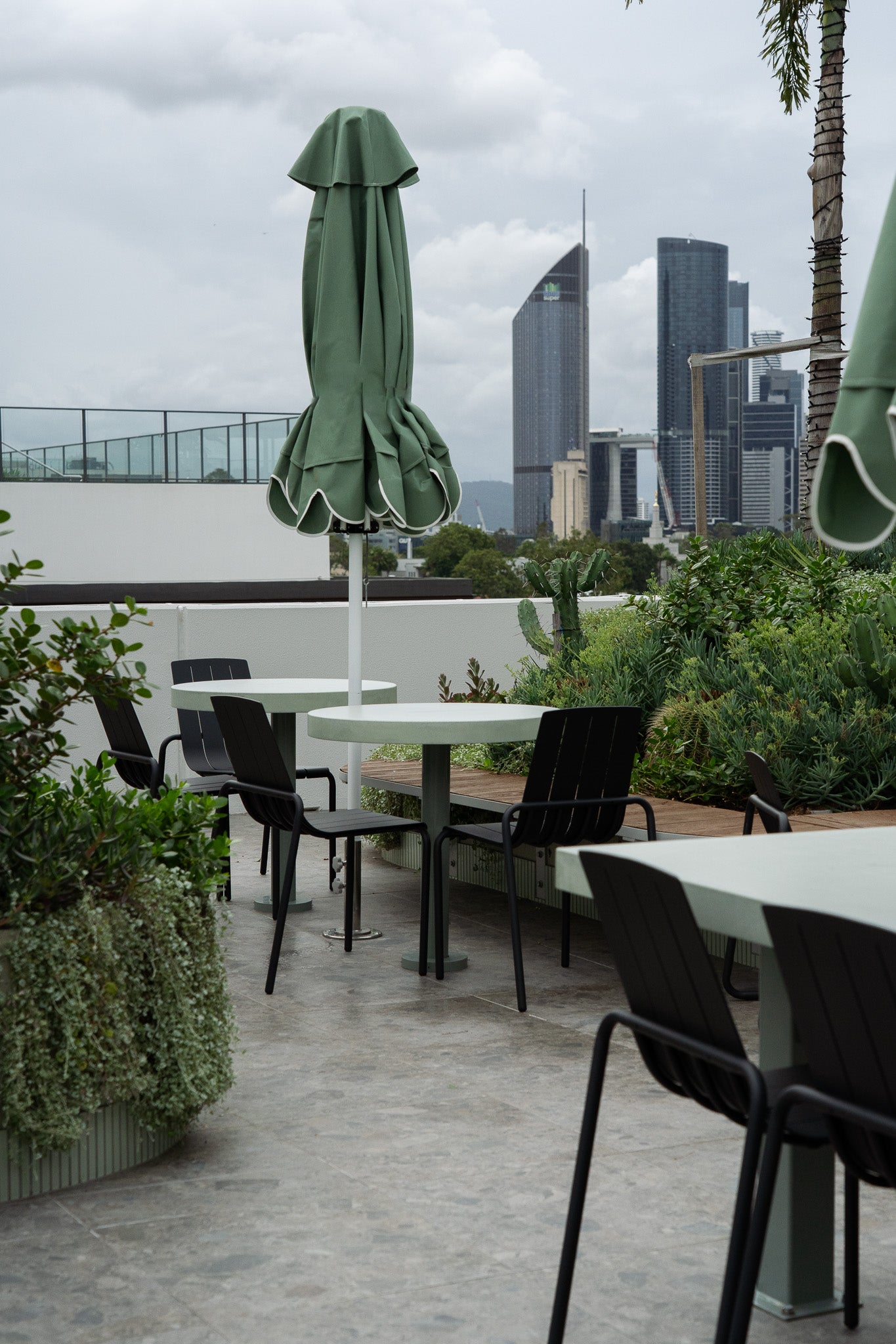An elevated outdoor terrace featuring custom green concrete tables with pedestal bases, black metal chairs, and closed green umbrellas. Surrounded by lush planter boxes and timber bench seating, the space overlooks Brisbane’s city skyline under a cloudy sky.