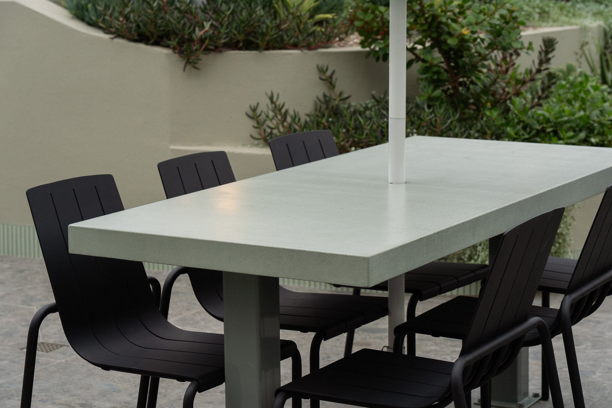 A rectangular green concrete outdoor dining table with a central umbrella hole, surrounded by six black metal chairs. The setting features lush greenery and soft beige rendered planter walls, creating a clean and contemporary alfresco environment.