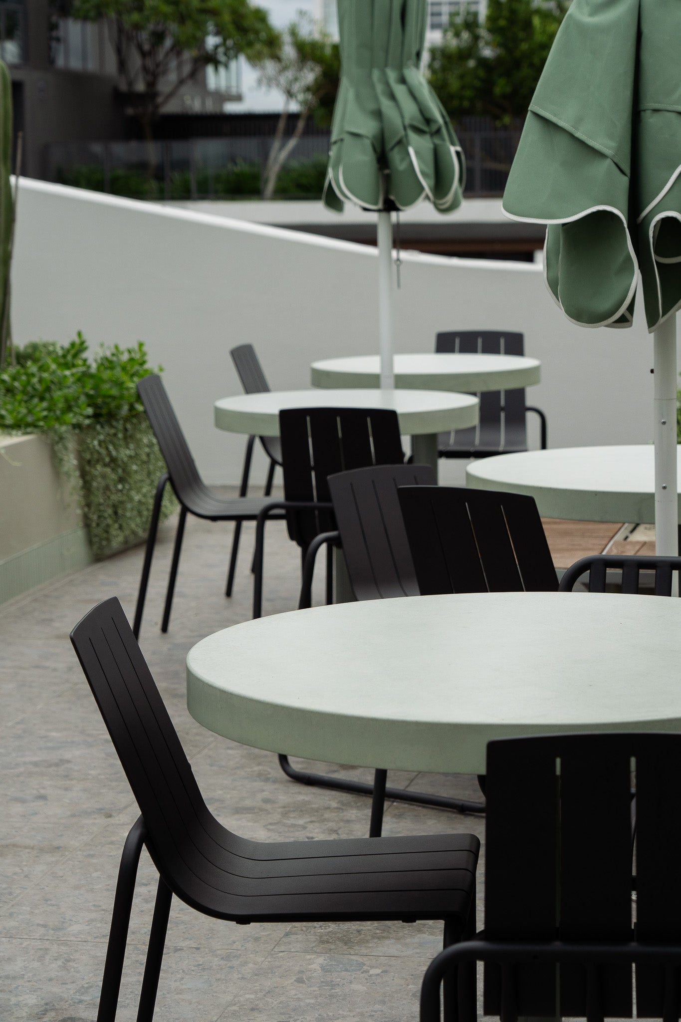 A row of round green concrete café tables with black outdoor chairs and closed green umbrellas, arranged on a tiled rooftop terrace. The space is framed by curved planters and minimalist architectural lines, creating a refined urban outdoor setting.