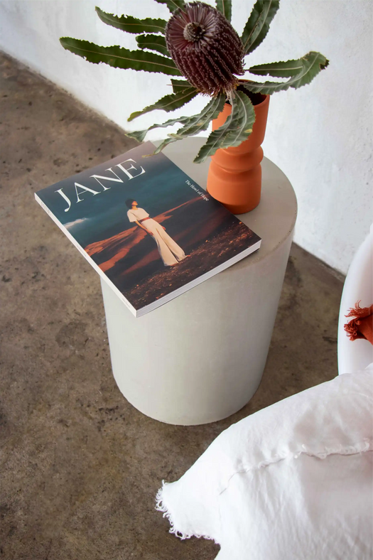 The Dune Concrete Side Table in a Mist Grey next to a modern white bubble chair with an issue of the French magazine Jane on top next to an orange vase.