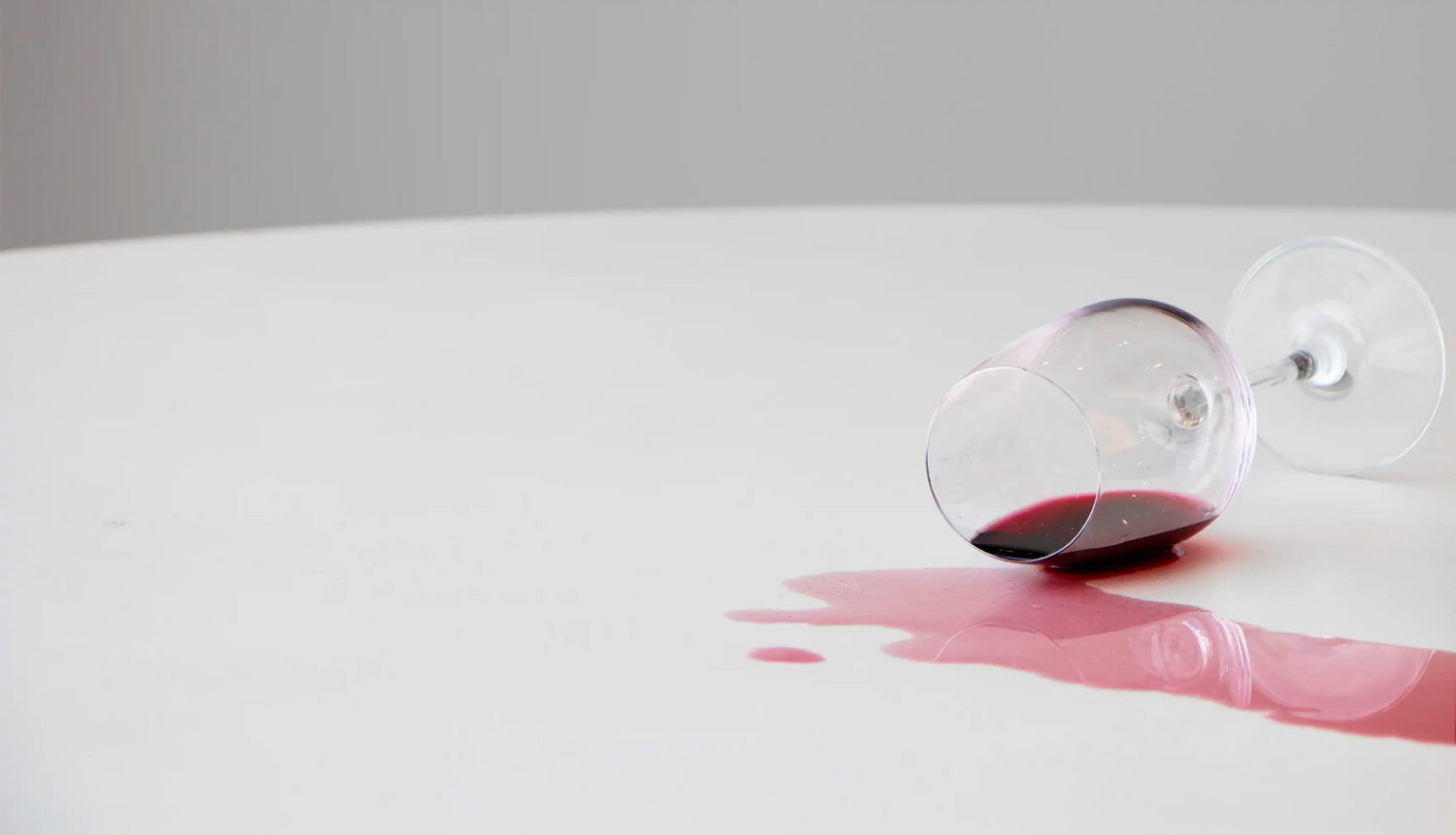 A wine glass tipped over spilling red wine on a white concrete benchtop.
