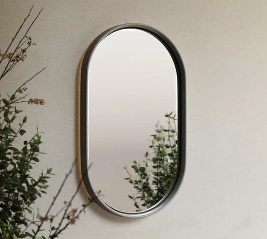 A grey Lunar Concrete Mirror on an outside beige wall with greenery on the side and in the reflection of the concrete mirror. 