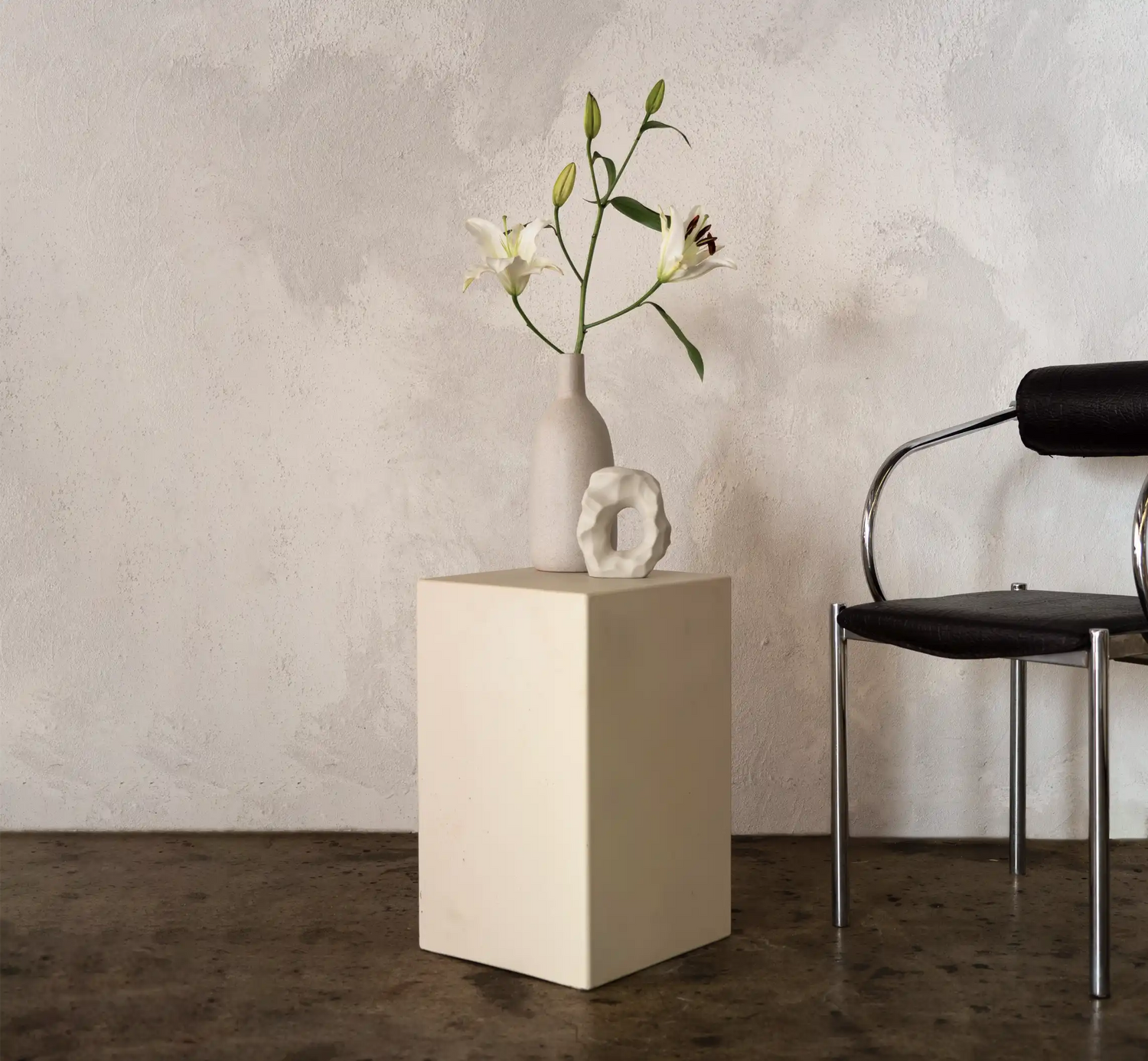 A Sand coloured Sahara Concrete Side Table in a minimalist living room next to a funky modern dining chair and a vase of lilies. 