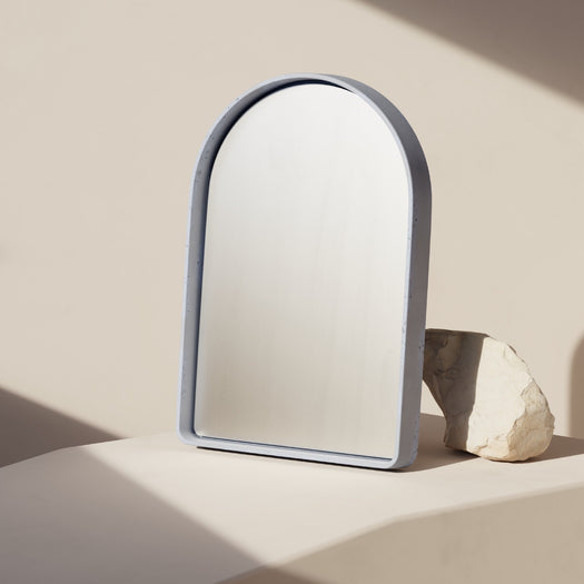 The Arc Concrete Mirror with a blue concrete frame leaning against a sand rock.