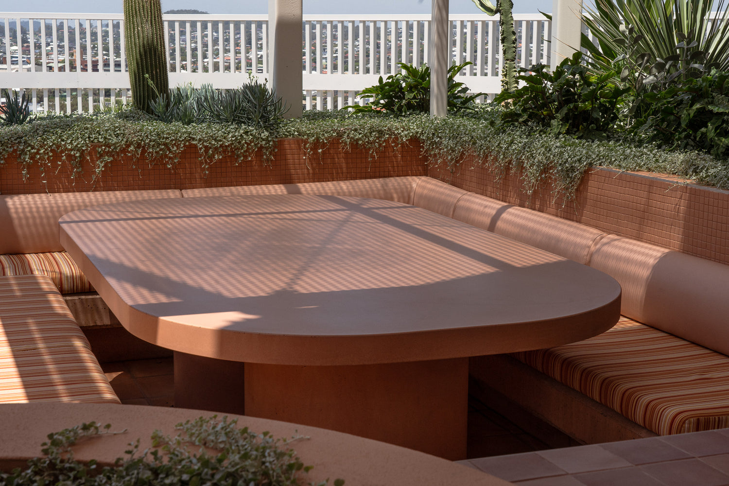 A custom terracotta-toned concrete table with rounded edges and a sturdy base, set within a sunken outdoor seating area. The table is surrounded by built-in cushioned benches upholstered in striped fabric, complementing the warm earthy hues.