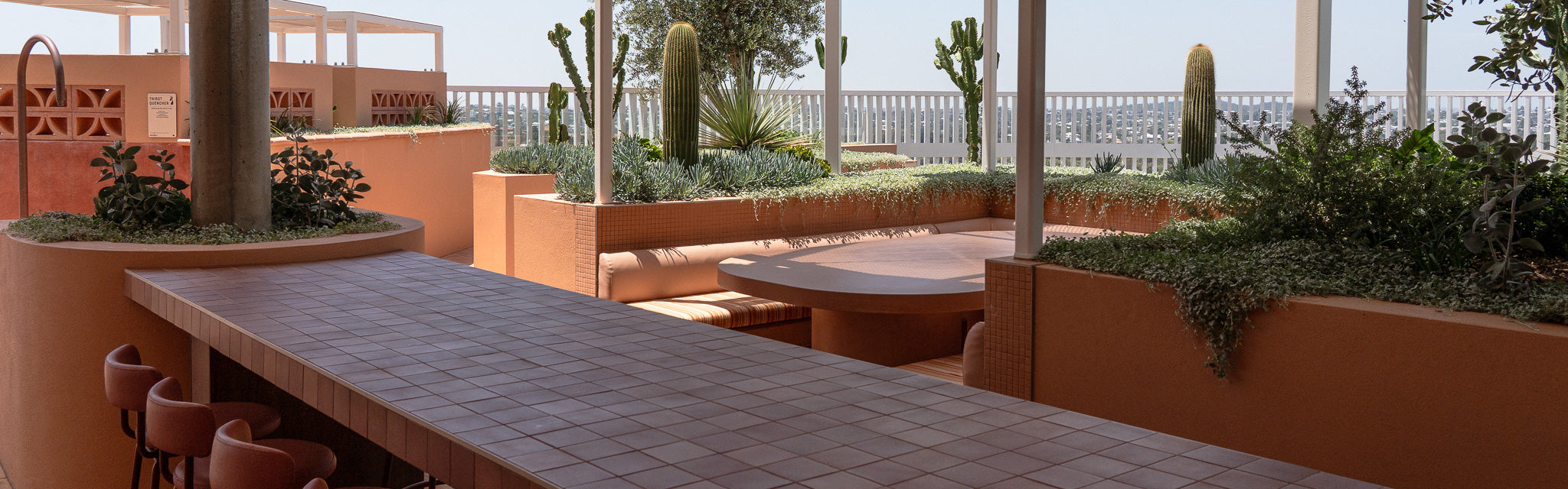 A beautifully designed outdoor terrace featuring earthy terracotta tones, lush greenery, and a blend of modern and natural elements. A custom concrete table sits  under a white canopy.
