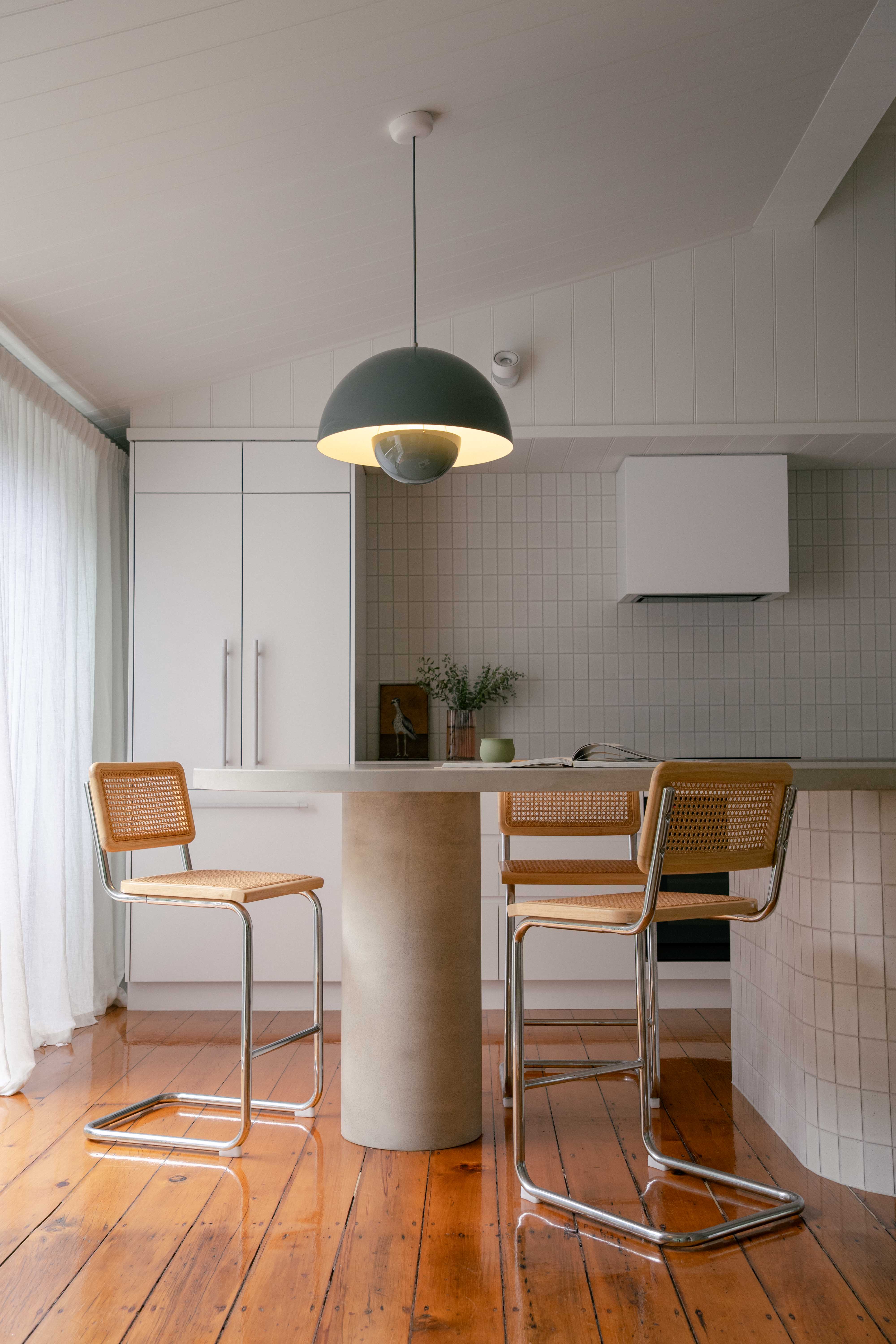 Custom concrete island kitchen bench with a rounded pillar support by Blend Concrete Design, featured in a bright, modern kitchen with timber floors, cane and chrome bar stools, and soft neutral finishes.