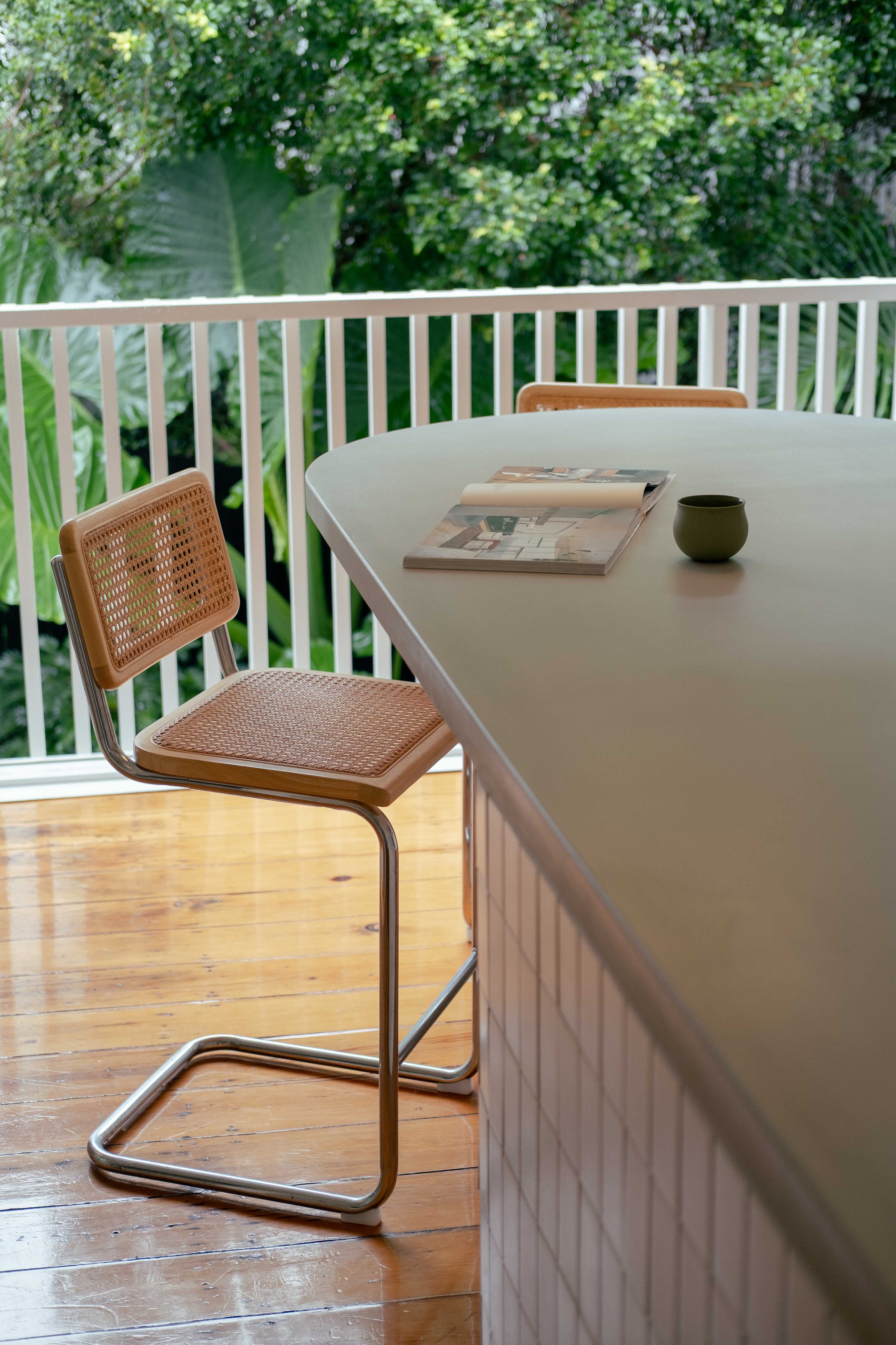 Custom concrete island kitchen bench crafted by Blend Concrete Design, featured in a light-filled space with timber floors, cane dining chairs, and lush greenery beyond the open balcony.